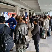 Eine Gruppe von Menschen steht in einem hellen Innenraum. Einige Personen sind mit Rucksäcken und Jacken bekleidet und stehen in einer Schlange vor einem Tisch. Im Hintergrund sind Poster und Informationsstände zu sehen, die eine Veranstaltung oder Messe anzeigen.