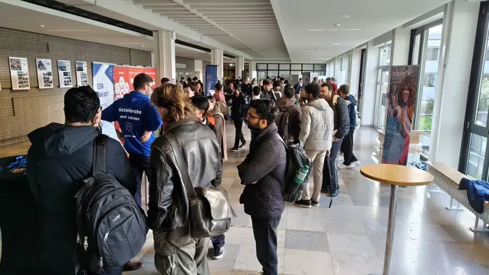 Eine Gruppe von Menschen steht in einem hellen Innenraum. Einige Personen sind mit Rucksäcken und Jacken bekleidet und stehen in einer Schlange vor einem Tisch. Im Hintergrund sind Poster und Informationsstände zu sehen, die eine Veranstaltung oder Messe anzeigen.