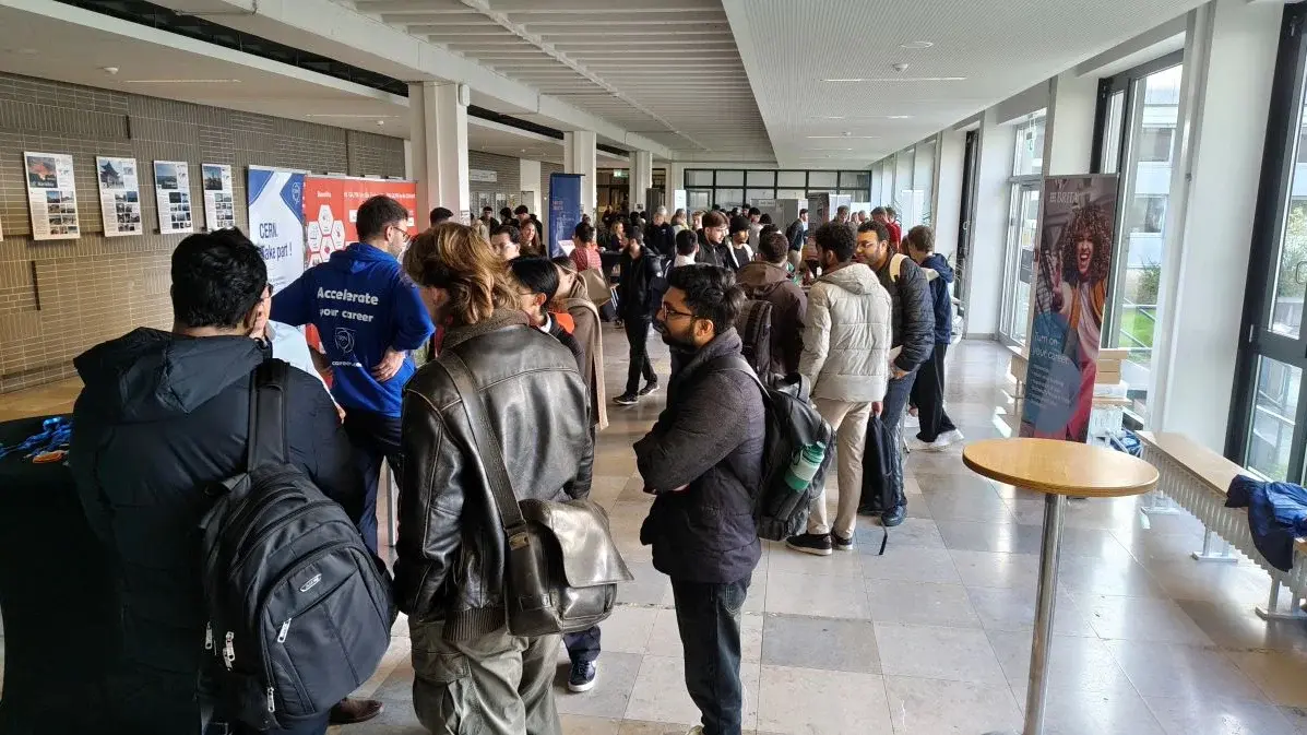 Eine Gruppe von Menschen steht in einem hellen Innenraum. Einige Personen sind mit Rucksäcken und Jacken bekleidet und stehen in einer Schlange vor einem Tisch. Im Hintergrund sind Poster und Informationsstände zu sehen, die eine Veranstaltung oder Messe anzeigen.