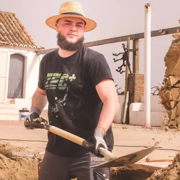 Ein junger Mann mit einem Hut und Bart steht an einer Baustelle und hält eine Schaufel. Er trägt ein schwarzes T-Shirt und Handschuhe und arbeitet im Sand. Im Hintergrund sind unscharfe Gebäude zu sehen.