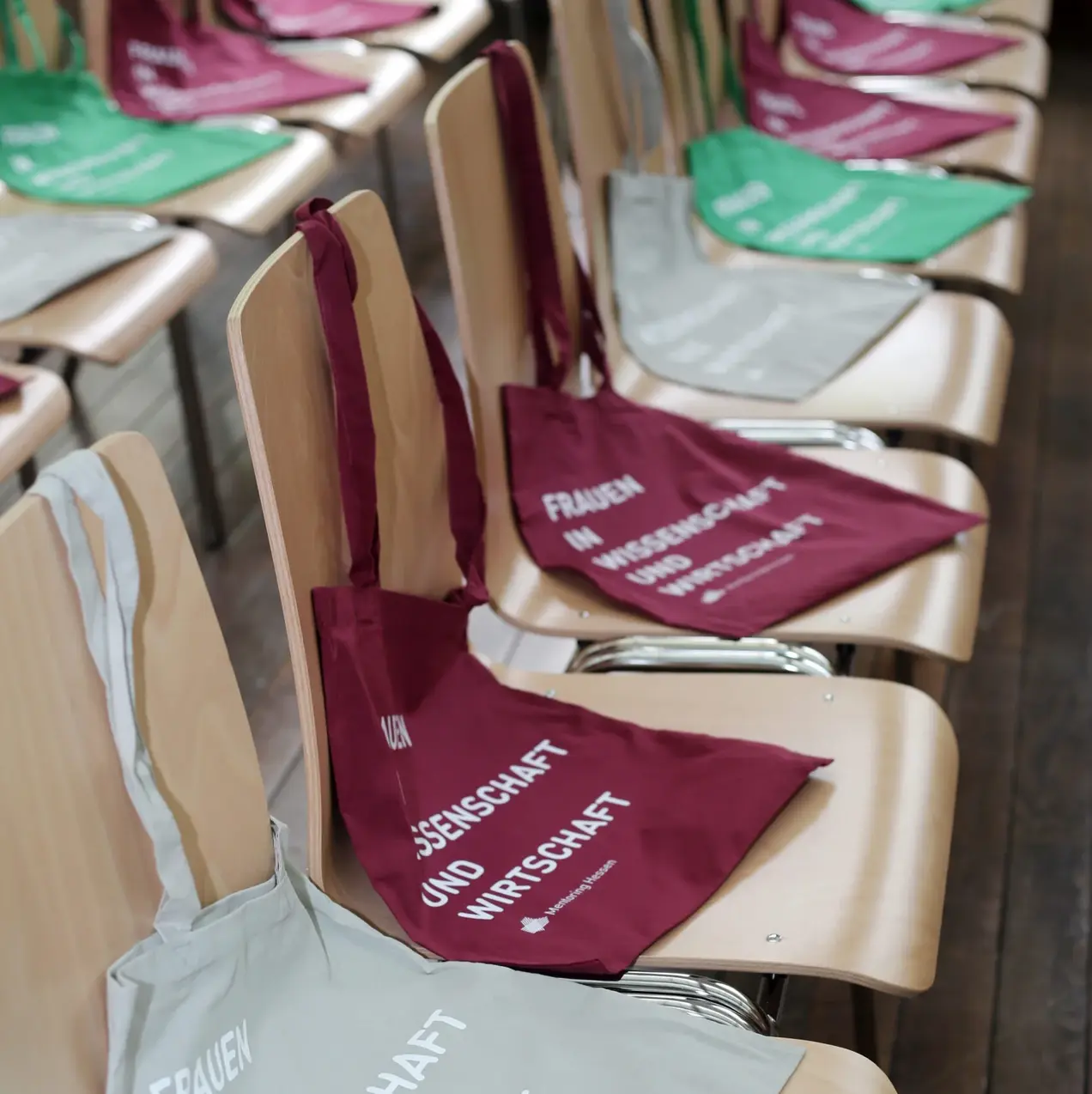 Auf den Stühlen liegen mehrfarbige Stofftaschen mit dem Schriftzug "Frauen in Wissenschaft und Wirtschaft". Die Taschen sind in verschiedenen Farben wie Grün, Rot und Grau und verdeutlichen das Thema der Veranstaltung.