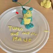 Eine bunt geformte Maus aus Knete sitzt auf einem weißen Teller. Unter der Maus stehen die Worte „Türen auf mit der MAUS“ in gelber Knete geschrieben. Im Hintergrund sind weitere Knetemängel sichtbar.