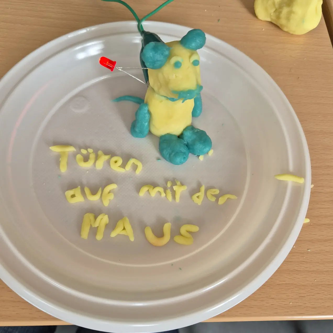 Eine bunt geformte Maus aus Knete sitzt auf einem weißen Teller. Unter der Maus stehen die Worte „Türen auf mit der MAUS“ in gelber Knete geschrieben. Im Hintergrund sind weitere Knetemängel sichtbar.