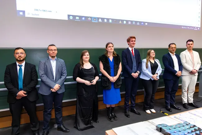 Eine Gruppe von acht Personen steht nebeneinander in einem Raum. Die Menschen tragen formelle Kleidung. Im Hintergrund ist eine Tafel sichtbar. Sie wirken freundlich und stehen in Erwartung, möglicherweise für eine Präsentation oder ein Event.