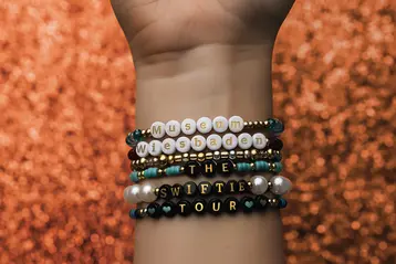 Auf einem Handgelenk sind mehrere bunte Armbänder sichtbar. Eines der Armbänder enthält die Aufschrift "Museum Wiesbaden", ein anderes "THE SWIFTIE TOUR". Die Armbänder sind aus verschiedenen Perlen und dekorativen Elementen gefertigt. Der Hintergrund ist glänzend und orangefarben.