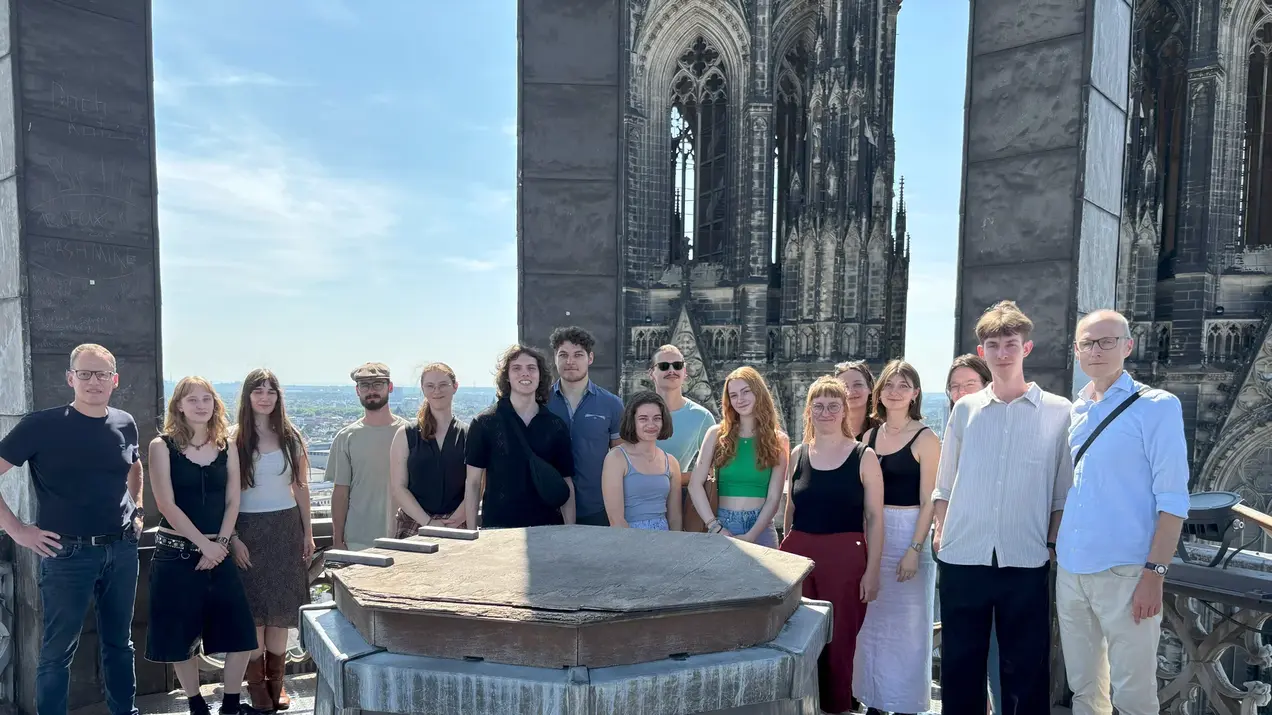 Baukulturerbe-Studierende auf dem Vierungsturm des Kölner Doms. Einige Personen stehen zusammen, während andere lächeln oder in die Kamera schauen. Im Hintergrund sind die Türme des Kölner Doms sichtbar und der Himmel ist klar und blau.