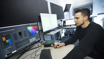 Ein junger Mann sitzt an einem Tisch vor zwei Monitoren, auf denen Farbgrafiken und Diagramme angezeigt werden. Er arbeitet konzentriert an einer Computertastatur, um audiovisuelle Daten oder Farbspektren zu analysieren oder zu bearbeiten. Im Hintergrund sind mehrere technische Geräte sichtbar.