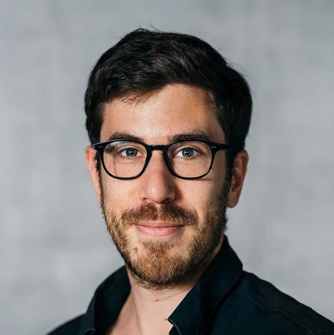 Ein junger Mann mit Brille und Bart lächelt in die Kamera. Er trägt ein schwarzes Hemd und hat kurzes, dunkles Haar. Der Hintergrund ist grau und unscharf, was den Fokus auf sein Gesicht legt.