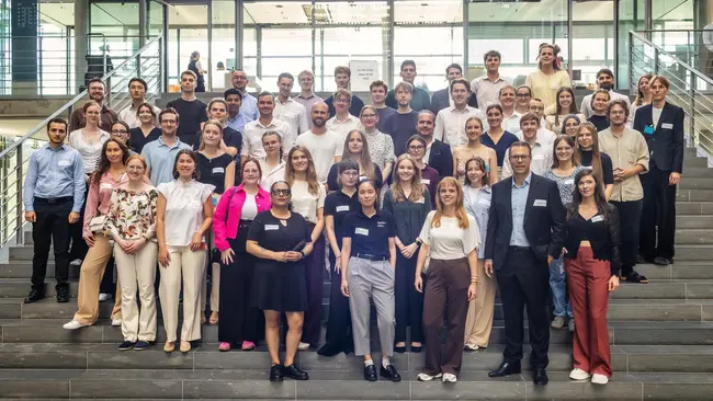 Eine Gruppe von etwa 50 Personen steht auf einer Treppe in einem modernen Gebäude. Die Teilnehmer haben unterschiedliche Kleidung und stehen gemeinsam für ein Foto, während sie lächeln. Namensschilder sind sichtbar. Die Veranstaltung scheint eine Konferenz oder ein Treffen zu sein.