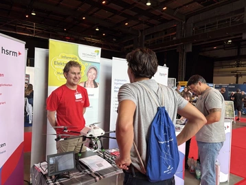 Auf der Messe steht ein Mann in einem roten T-Shirt mit dem Logo einer Hochschule vor einem Stand. Er spricht mit zwei Besuchern, einer davon hält eine Drohne in den Händen. Im Hintergrund sind weitere Informationsstände zu sehen.