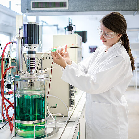 Studentin in Laborausrüstung spritzt eine grüne Flüssgkeit in einen Bioreaktor