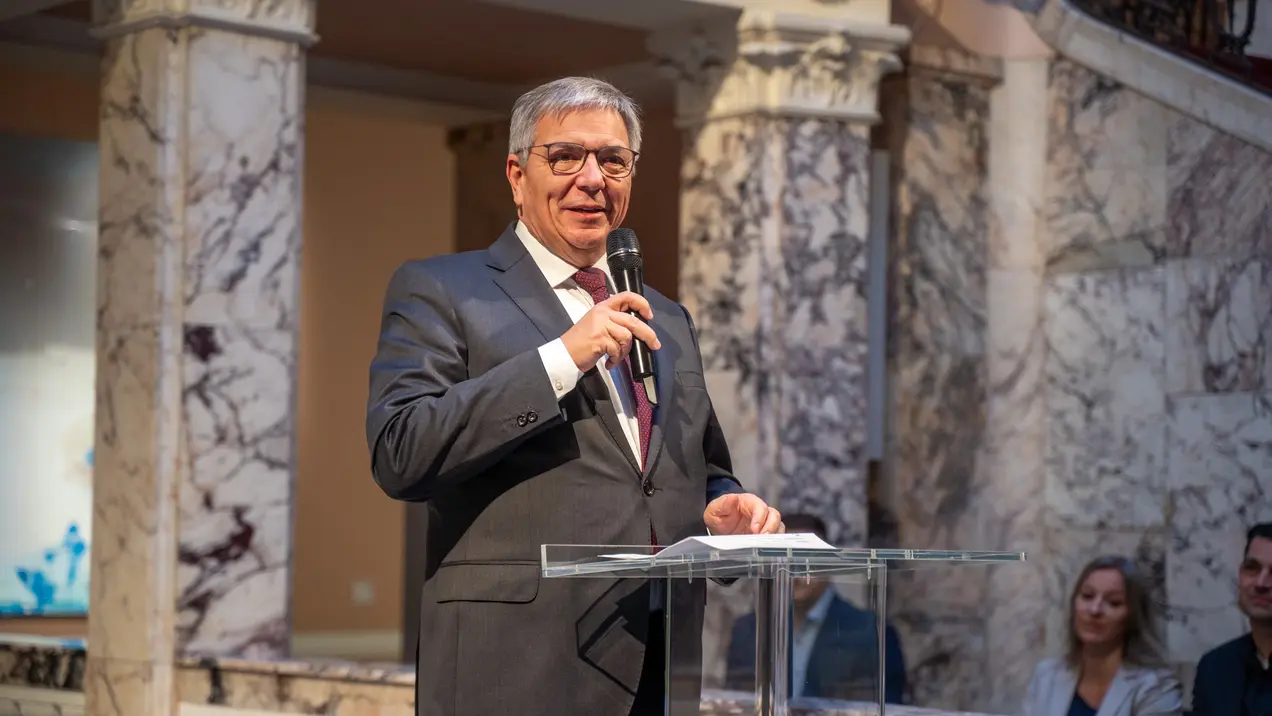 Ein Mann in Anzug steht am Rednerpult und hält ein Mikrofon. Er spricht in einem festlichen Raum mit Marmorwänden, während einige Zuhörer im Hintergrund aufmerksam zuhören.