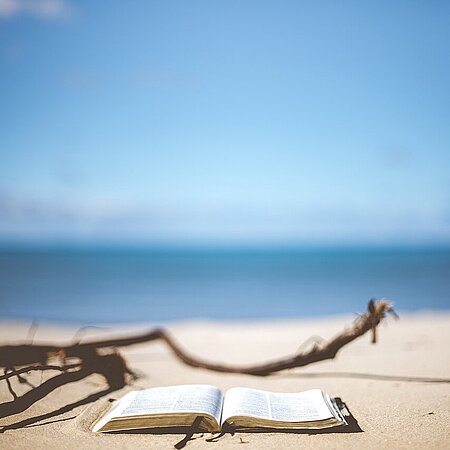 Ein aufgeschlagenes Buch liegt am Strand. Man kann das Meer im Hintergrund sehen