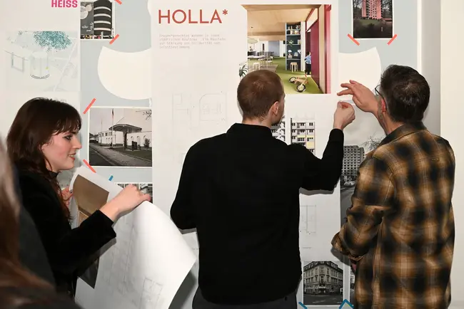 Drei Personen stehen vor einer Plänen der Bachelor- und Master-Thesisarbeiten des Studiengangs Architektur. Die Männer zeigen auf bestimmte Bereiche, während eine Frau einen Plan in der Hand hält. Die Wand enthält Informationen zu einem architektonischen Projekt.