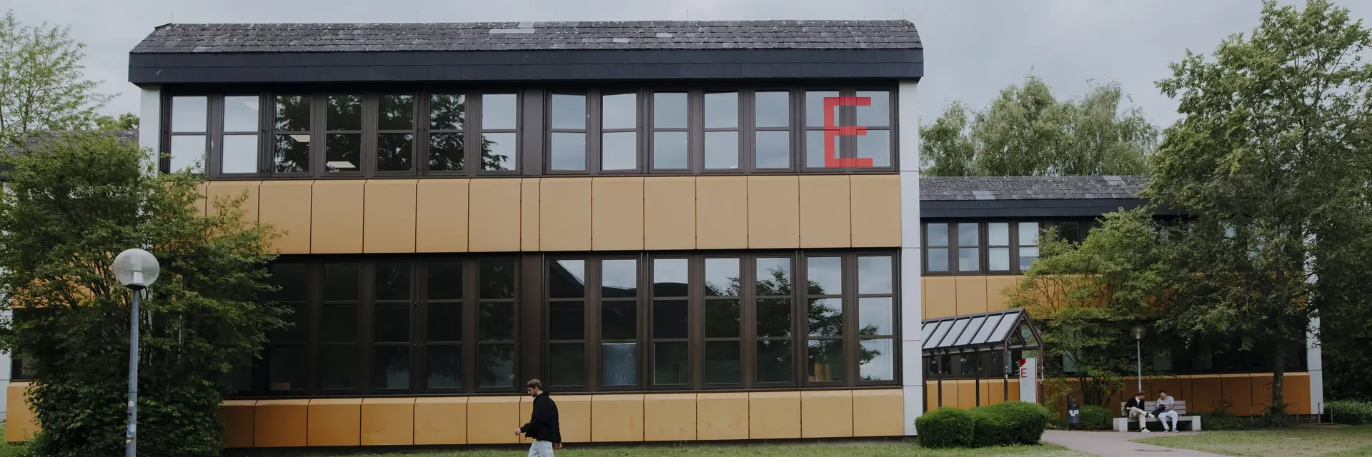 Ein modernes Gebäude mit großen Fenstern, dessen Fassade teilweiße in Gelb gestaltet ist. Vor dem Gebäude steht eine Person mit schwarzem Oberteil und hellen Hosen. Auf dem Rasen wachsen einige Bäume und Büsche.