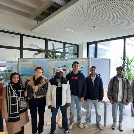 A diverse group of nine people stands together indoors, smiling at the camera. They are dressed in casual clothing, with large windows in the background showing greenery outside. The atmosphere appears friendly and welcoming.