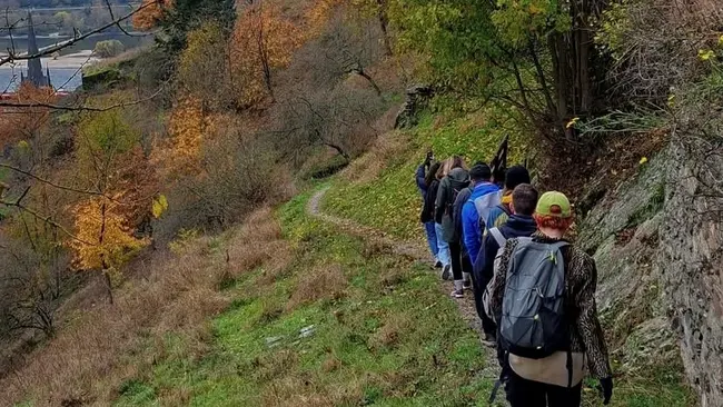 Eine Gruppe von Wanderern bewegt sich auf einem schmalen Weg entlang eines Hangs. Die Umgebung ist herbstlich mit bunten Blättern. Im Hintergrund ist eine Landschaft mit Wasser sichtbar. Die Wanderer tragen Rucksäcke und sind in Winterkleidung.