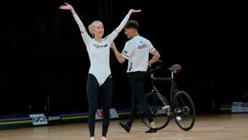 Die Aufnahme zeigt zwei Sportler auf einer Wettkampfbühne. Eine Athletin in einem weißen Anzug hebt die Arme in einer freudigen Geste, während ein männlicher Sportler im Hintergrund auf einem Fahrrad steht. Beide zeigen ihre sportlichen Fähigkeiten bei einem Wettbewerb.