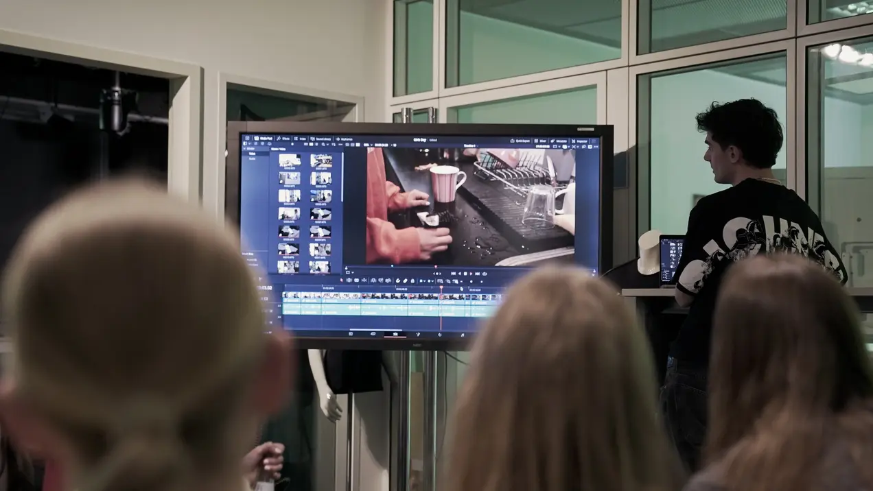 Eine Gruppe von Personen sitzt und beobachtet einen Bildschirm, auf dem ein Videobearbeitungsprogramm geöffnet ist. Der Bildschirm zeigt Hände, die Tassen und andere Objekte bearbeiten. Im Hintergrund steht eine Person, die die anderen anleitet oder präsentiert.