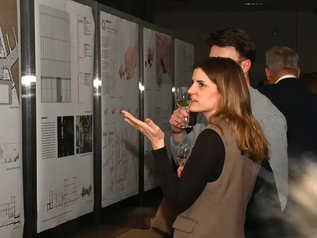 Eine Studentin hält ein Glas Sekt und zeigt mit der Hand auf Präsentationen des Bachelor und Master Architektur an einer Wand. Sie diskutiert mit einem Mann neben ihr, während im Hintergrund weitere Anwesende und Informationen sichtbar sind. 