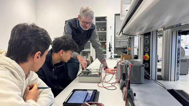 Ein Lehrer zeigt zwei Schülern an einem Tisch, wie man Messgeräte verwendet. Die Schüler, einer in einem weißen Hoodie und der andere in schwarzem Pullover, beobachten aufmerksam. Auf dem Tisch sind verschiedene elektrische Geräte und ein Tablet sichtbar, das zur Dokumentation verwendet wird.