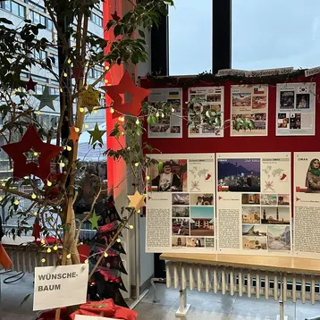 Der Wünschtbaum steht in einem Raum. An ihm hängen rote Sterne und ein Schild mit der Aufschrift „WÜNSCHE-BAUM“ sowie „Jeder Stern = 1 Wunsch“. Im Hintergrund sind Informationsplakate und Bilder ausgestellt, die wahrscheinlich die Veranstaltung oder Initiative näher erläutern.