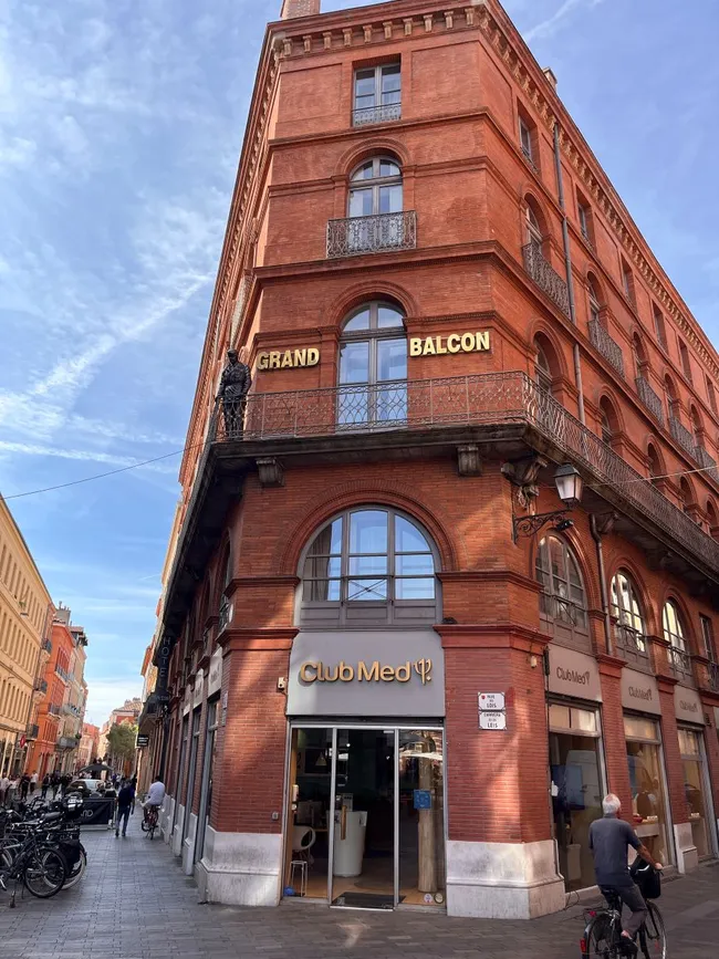 Ein rotes Ziegelgebäude an einer Straßenecke mit den Schriftzügen „GRAND BALCON“ und „Club Med“. Der Eingangsbereich von Club Med ist sichtbar, dazu eine belebte Straße mit Parkplätzen und Fahrrädern. Der Himmel ist blau mit wenigen Wolken.
