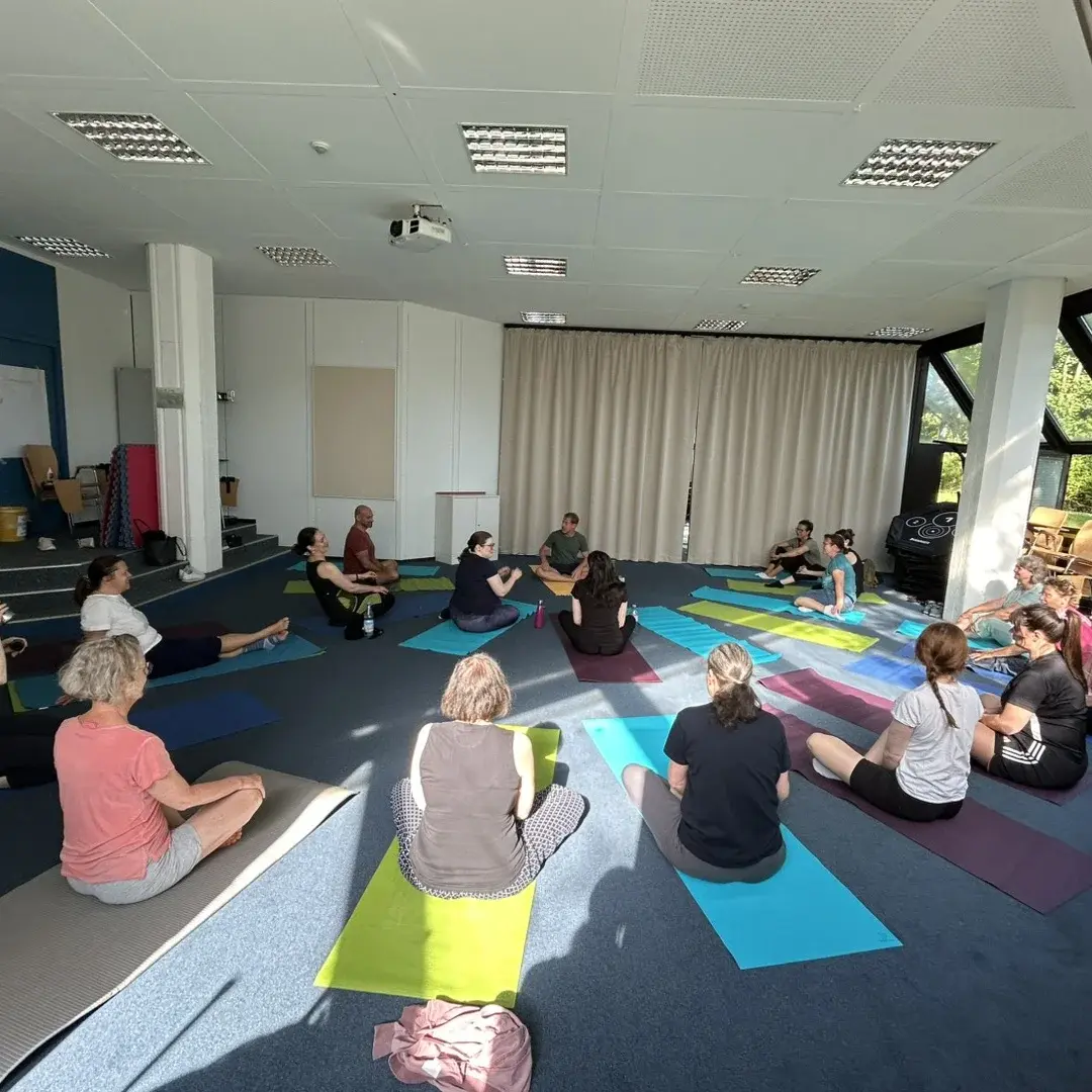 In einem hellen Raum sitzen etwa 15 Personen auf Yogamatten. Sie arbeiten gemeinsam in einer Gruppenübung. Einige blicken zum Lehrer in der Mitte, während andere entspannt auf den Matten sitzen. Durch große Fenster strömt Tageslicht in den Raum.