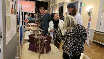 Interessierte Personen stehen vor einem Architekturmodell, ein Student erklärt seinen Entwurf. Im Hintergrund sind weitere Modelle und Pläne von anderen Entwürfen zu sehen.