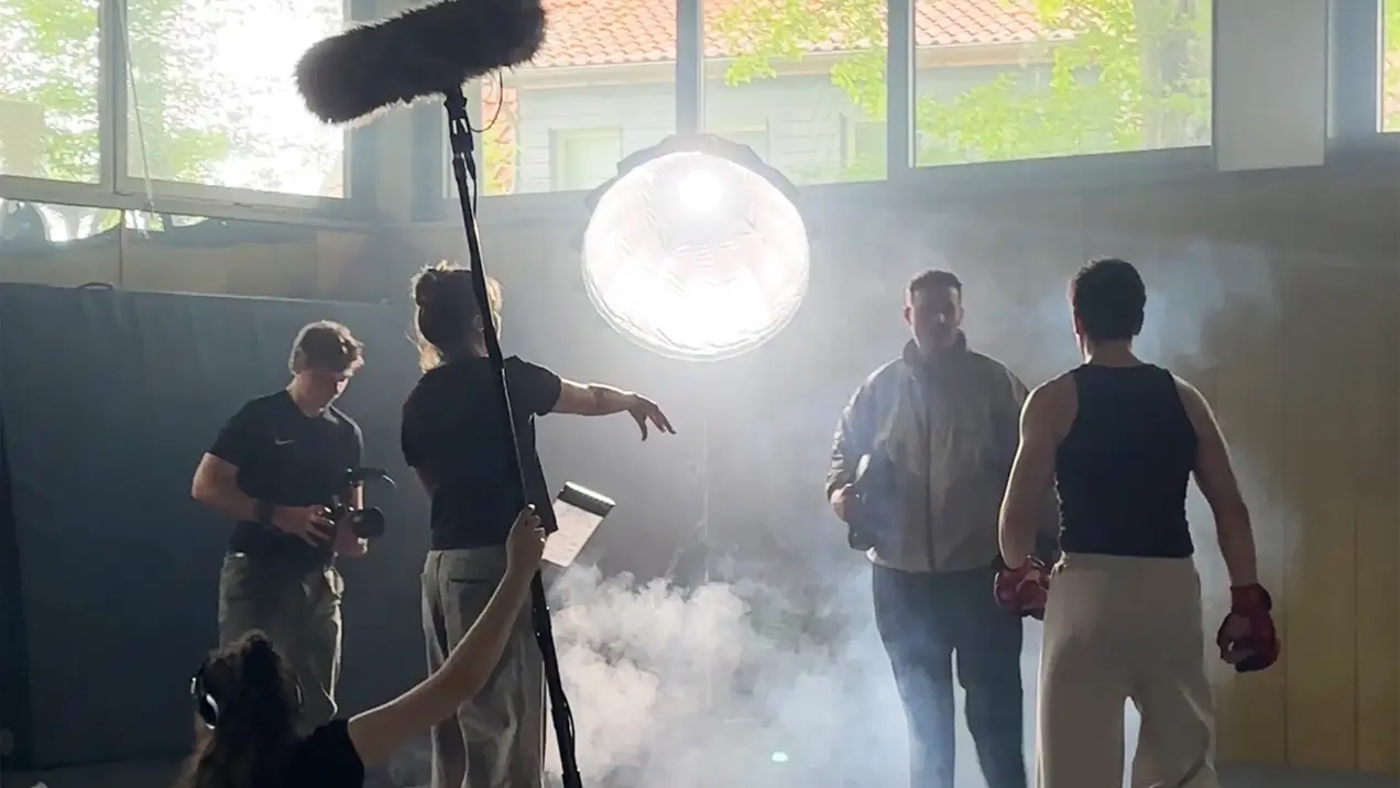 Ein Drehset mit mehreren Personen, darunter ein Filmemacher und ein Schauspieler in Boxkleidung. Im Hintergrund ist ein großes Lichtgerät zu sehen, während Nebel im Raum verteilt ist. Eine Person hält ein Mikrofon. Die Atmosphäre ist dynamisch und filmisch.