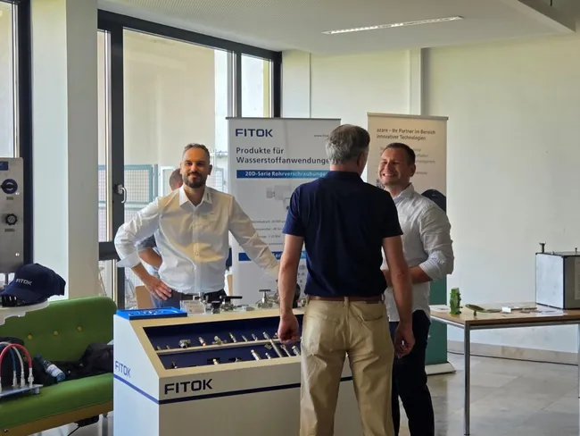 Drei Männer stehen an einem Stand von FITOK, der Produkte für Wasserstoffanwendungen präsentiert. Sie unterhalten sich freundlich. Im Hintergrund sind Werbebanner sichtbar, die Informationen über das Unternehmen enthalten.