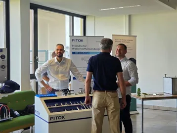Drei Männer stehen an einem Stand von FITOK, der Produkte für Wasserstoffanwendungen präsentiert. Sie unterhalten sich freundlich. Im Hintergrund sind Werbebanner sichtbar, die Informationen über das Unternehmen enthalten.