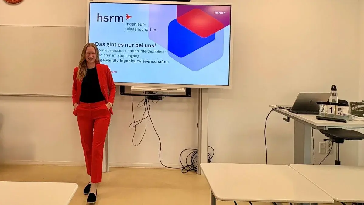 Eine Frau in einem roten Anzug steht selbstbewusst vor einem Bildschirm in einem Klassenraum. Auf dem Bildschirm steht der Text „hsrm - Ingenieurwissenschaften“ und „Das gibt es nur bei uns!“, der Informationen über Ingenieurstudiengänge präsentiert.