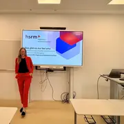 Eine Frau in einem roten Anzug steht selbstbewusst vor einem Bildschirm in einem Klassenraum. Auf dem Bildschirm steht der Text „hsrm - Ingenieurwissenschaften“ und „Das gibt es nur bei uns!“, der Informationen über Ingenieurstudiengänge präsentiert.