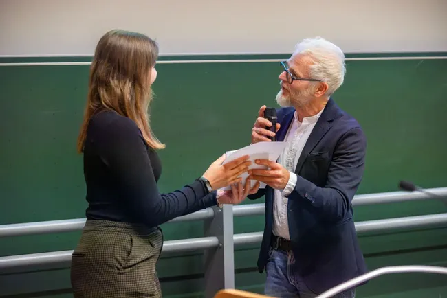 Eine Frau und ein älterer Mann stehen in einem akademischen Umfeld. Die Frau hält ein Dokument in der Hand, während der Mann mit einem Mikrofon spricht. Beide scheinen an einem Gespräch oder einer offiziellen Übergabe beteiligt zu sein.