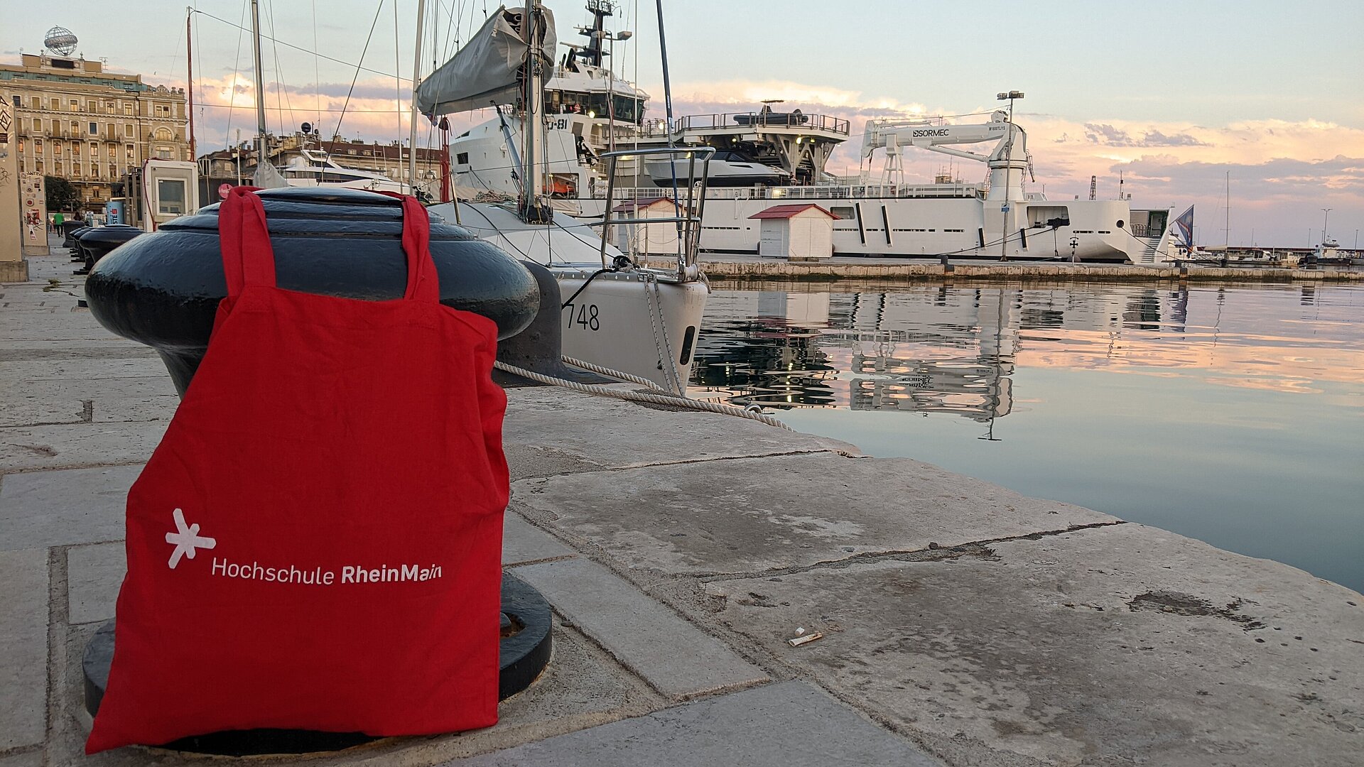 Die rote HSRM Tasche lehnt an einem Poller im Hafen von Rijeka, im Hintergrund das Meer und ein Segelboot
