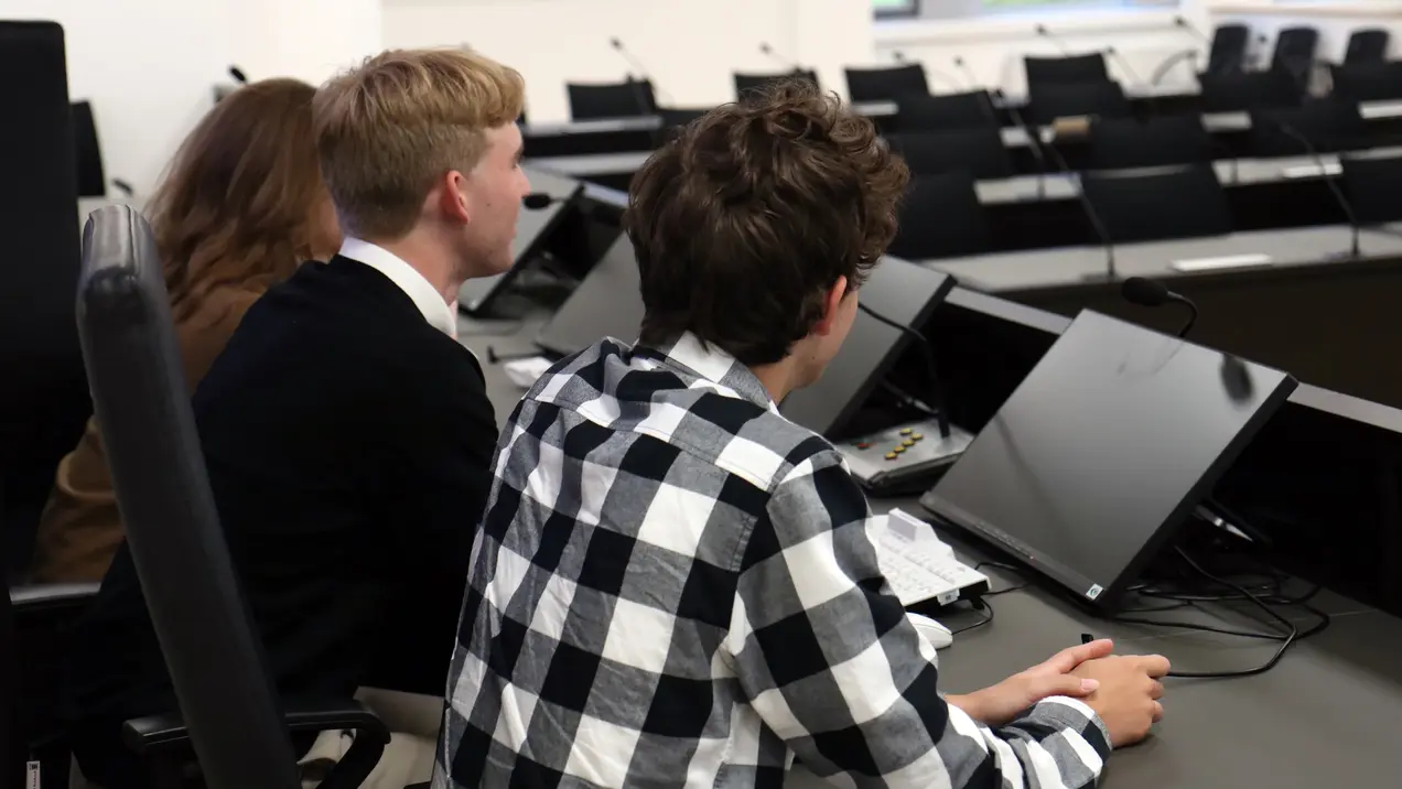 Drei junge Menschen sitzen an einem Tisch in einem modernen Raum mit Laptops. Sie blicken aufmerksam nach vorne, während sie an einer Diskussion oder Präsentation teilnehmen. Die Anordnung der Laptops deutet auf ein formelles Meeting hin.