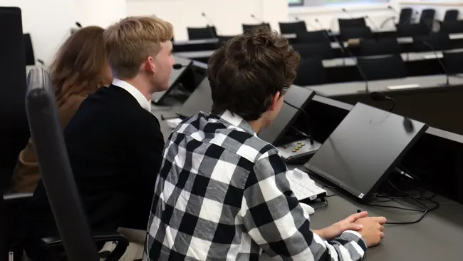 Drei junge Menschen sitzen an einem Tisch in einem modernen Raum mit Laptops. Sie blicken aufmerksam nach vorne, während sie an einer Diskussion oder Präsentation teilnehmen. Die Anordnung der Laptops deutet auf ein formelles Meeting hin.