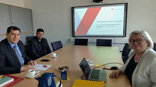 Drei Personen sitzen an einem Konferenztisch in einem Besprechungsraum. Auf einem großen Bildschirm steht eine Präsentation mit dem Titel „Die Förderung der persönlichen Weiterbildung“. Auf dem Tisch stehen Getränke und Unterlagen.