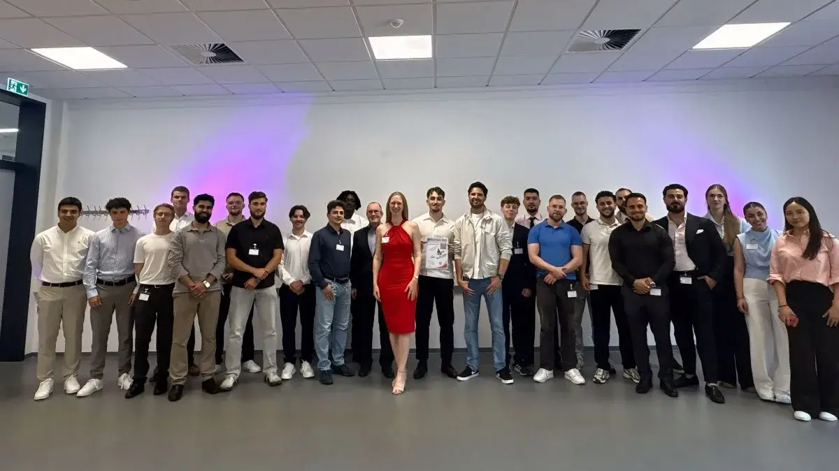 Eine Gruppe von etwa 25 Personen steht in einem Raum mit neutralem Hintergrund. Im Vordergrund steht eine Frau in einem roten Kleid, umgeben von Männern und Frauen in verschiedenen Kleidungsstilen. Alle Teilnehmer lächeln und scheinen an einem Event oder einer Feier teilzunehmen.