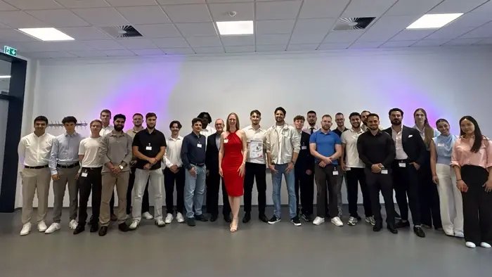 Eine Gruppe von etwa 25 Personen steht in einem Raum mit neutralem Hintergrund. Im Vordergrund steht eine Frau in einem roten Kleid, umgeben von Männern und Frauen in verschiedenen Kleidungsstilen. Alle Teilnehmer lächeln und scheinen an einem Event oder einer Feier teilzunehmen.