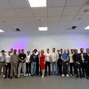 Eine Gruppe von etwa 25 Personen steht in einem Raum mit neutralem Hintergrund. Im Vordergrund steht eine Frau in einem roten Kleid, umgeben von Männern und Frauen in verschiedenen Kleidungsstilen. Alle Teilnehmer lächeln und scheinen an einem Event oder einer Feier teilzunehmen.