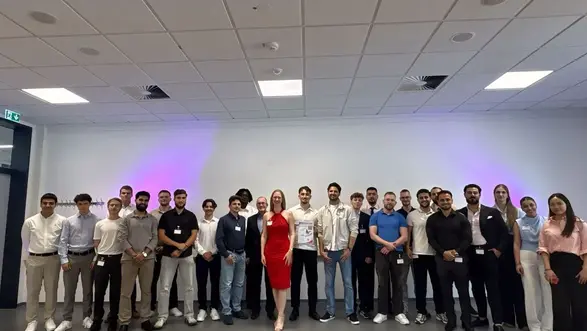 Eine Gruppe von etwa 25 Personen steht in einem Raum mit neutralem Hintergrund. Im Vordergrund steht eine Frau in einem roten Kleid, umgeben von Männern und Frauen in verschiedenen Kleidungsstilen. Alle Teilnehmer lächeln und scheinen an einem Event oder einer Feier teilzunehmen.