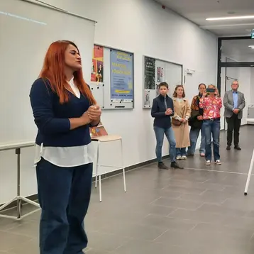 Eine Person mit roten Haaren steht vorne und spricht zu einer Gruppe von Menschen, die im Hintergrund stehen. Der Raum ist hell erleuchtet und hat eine moderne Atmosphäre. An einer Wand sind mehrere Plakate sichtbar.