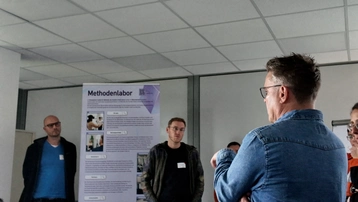 Eine Gruppe von Personen steht in einem Raum. Im Hintergrund ist ein Plakat mit dem Titel "Methodenlabor" sichtbar. Eine Person spricht, während die anderen aufmerksam zuhören. Das Bild zeigt eine Diskussion oder Präsentation.