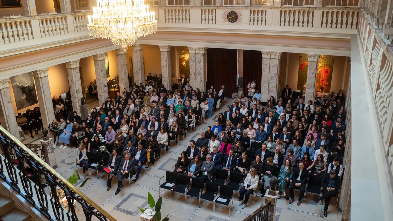 Eine große Versammlung findet in einem eleganten Saal statt, in dem viele Menschen in Reihen sitzen. Die Anordnung der Stühle ist geordnet, und eine beeindruckende Kristallleuchte hängt von der Decke. Die Atmosphäre wirkt formal und konzentriert.