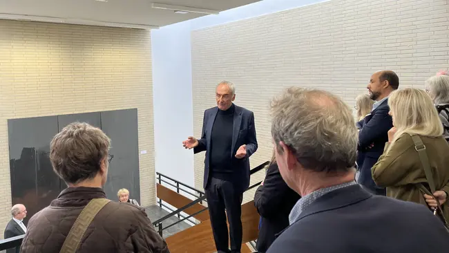 Ein Mann steht auf einer Treppe und spricht zu einer Gruppe von Menschen, die ihm zuhören. Die Umgebung ist modern und hell, mit einer klaren architektonischen Gestaltung im Hintergrund.