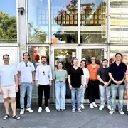 Eine Gruppe von elf Personen steht vor einem Gebäude. Sie lächeln und tragen lässige Kleidung. Hinter ihnen ist eine große Glasfront mit reflektierenden Fenstern zu sehen. Es ist ein sonniger Tag, und die Atmosphäre wirkt freundlich und einladend.