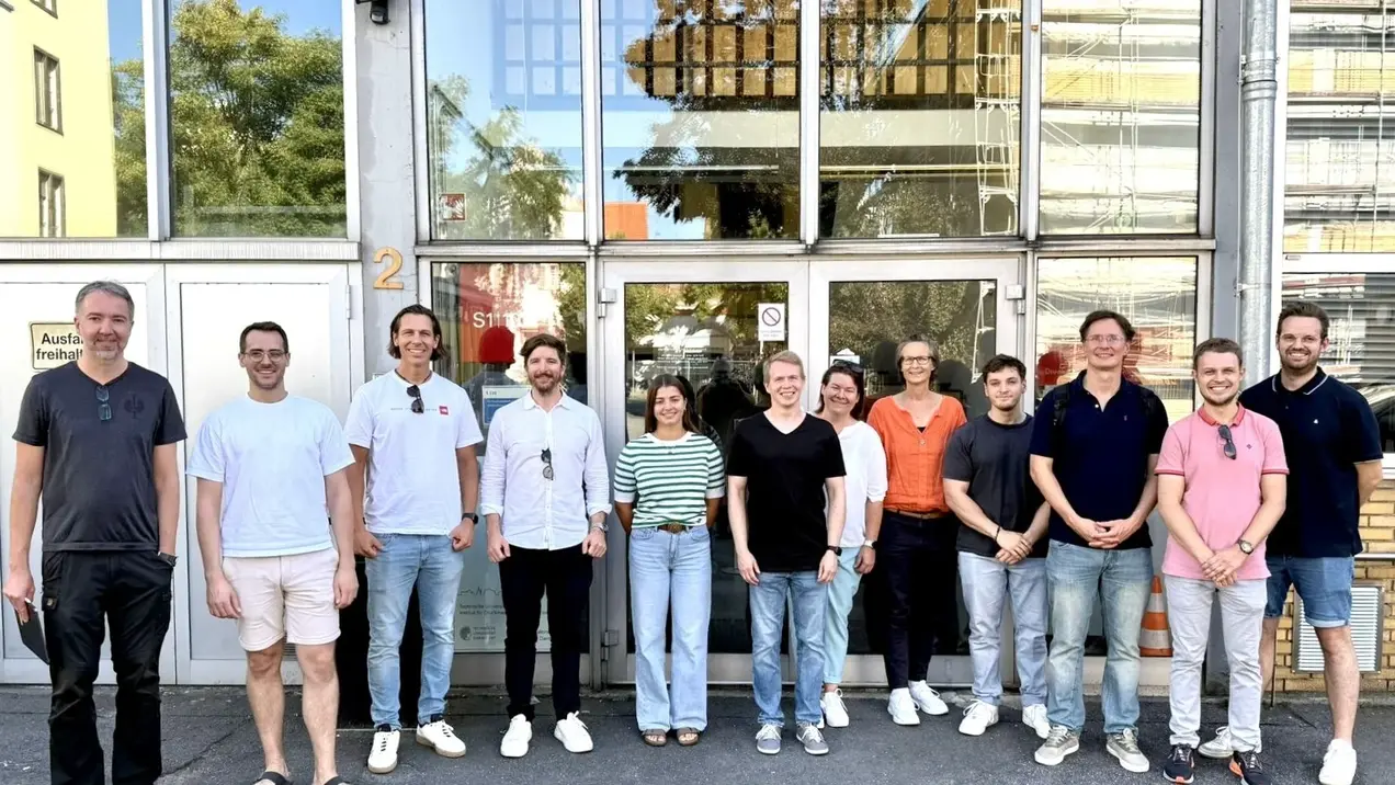 Eine Gruppe von elf Personen steht vor einem Gebäude. Sie lächeln und tragen lässige Kleidung. Hinter ihnen ist eine große Glasfront mit reflektierenden Fenstern zu sehen. Es ist ein sonniger Tag, und die Atmosphäre wirkt freundlich und einladend.