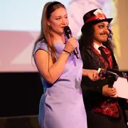Moderatorin Lilly trägt ein blaues Kleid und hält ein Mikrofon in der Hand, ihr Kollege Joel steht verkleidet als Tonmacher neben ihr
